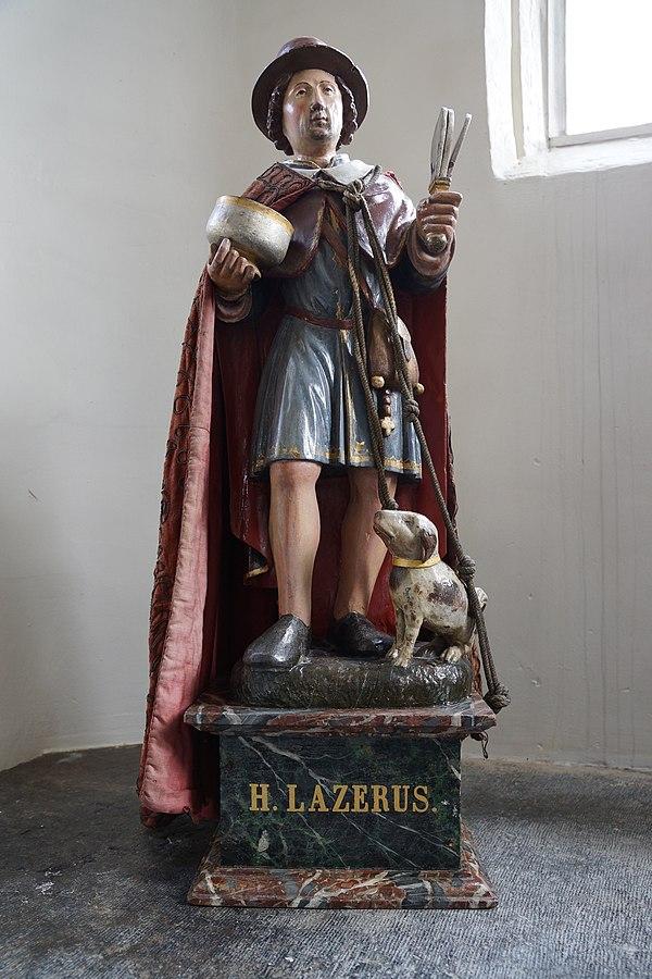 Foto van het beeld van de heilige Lazarus. Hij heeft een hoed op en mantel aan. En een hond aan de leiband. Op zijn voetstuk staat: "H. Lazarus".